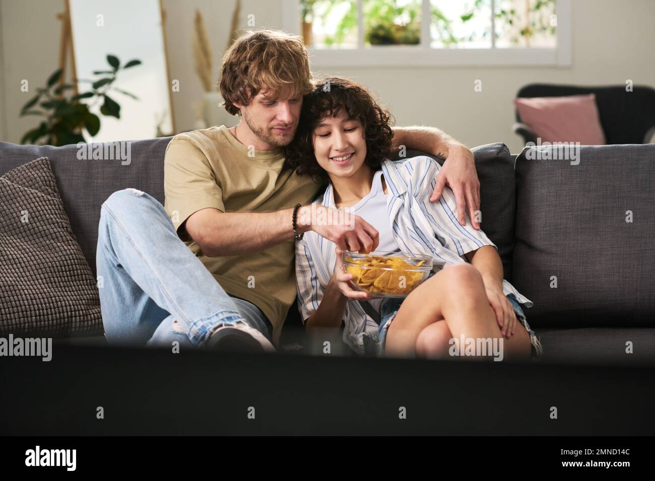 Jeune homme prenant des chips de pomme de terre dans un bol tenu par sa femme tout en se relaxant sur un canapé confortable et en regardant quelque chose à la télévision Banque D'Images