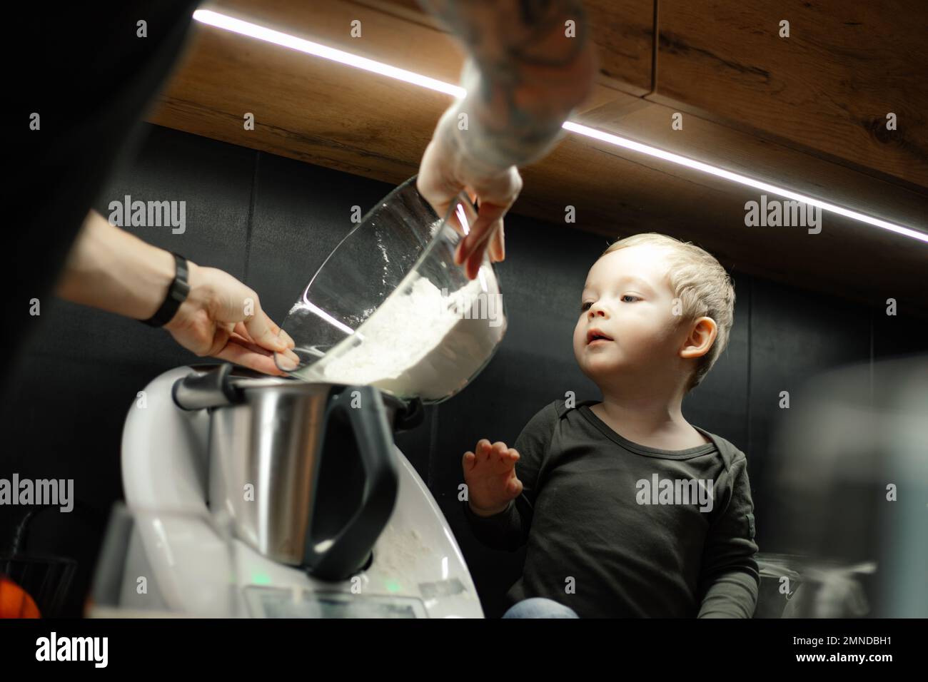 Versez la farine du bol en verre profond dans le blender. Un petit garçon aide le papa à préparer le dîner. Bébé dans la cuisine parmi les appareils modernes. Banque D'Images