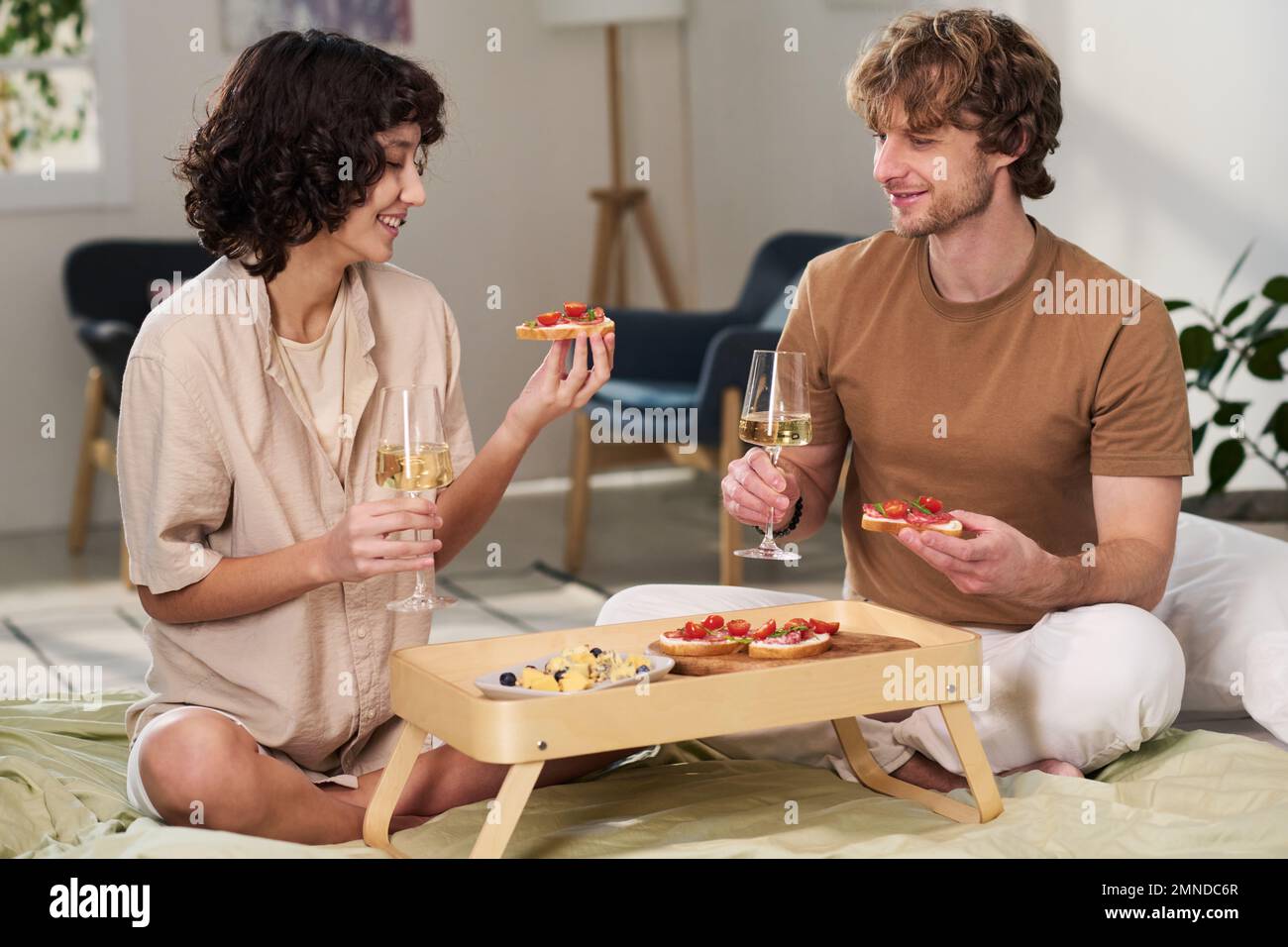 Une jeune femme heureuse et son mari qui tiennent des sandwiches et des flûtes de champagne tout en étant assis au lit et en prenant le petit déjeuner Banque D'Images