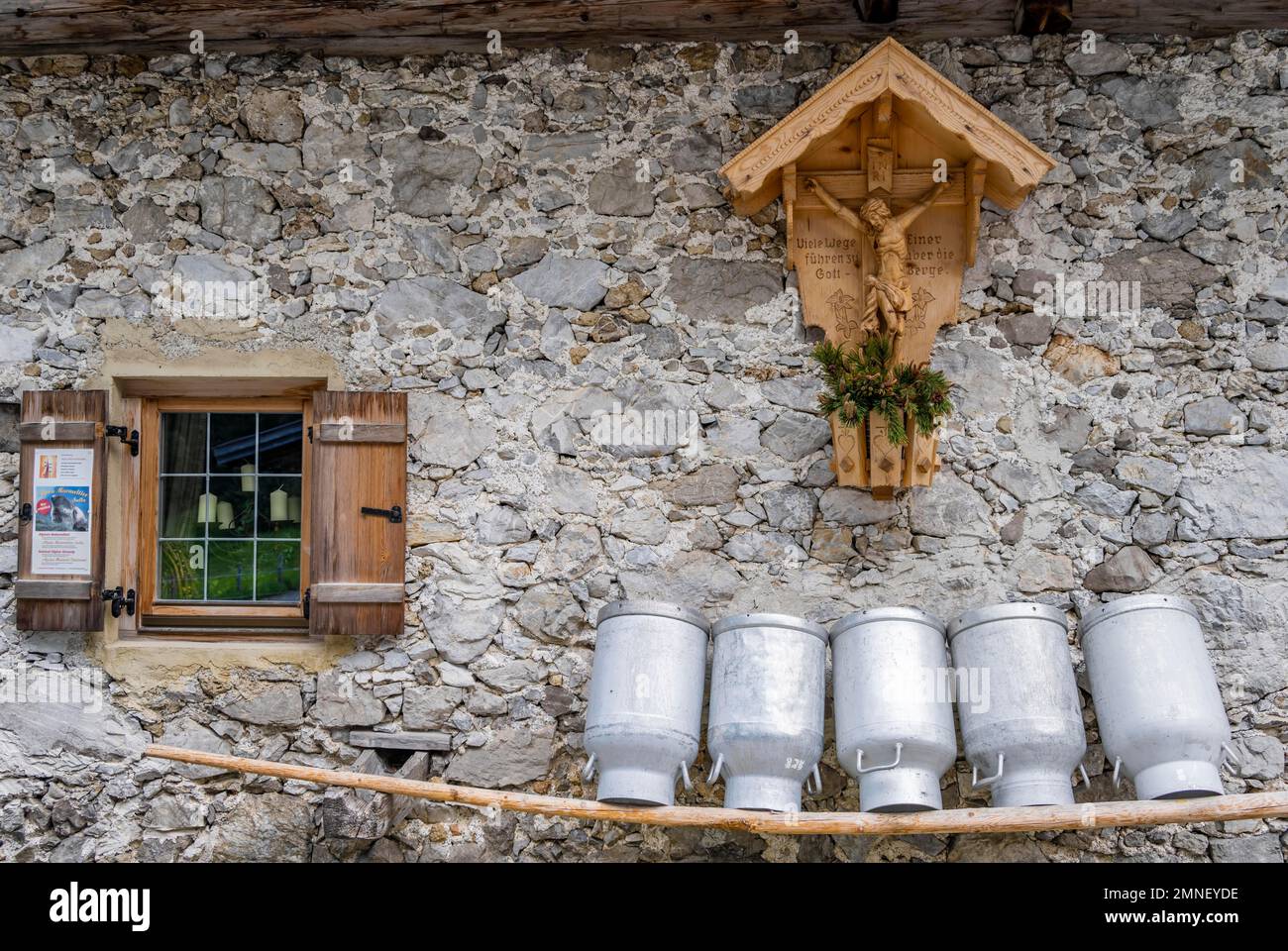 Canettes de lait alignées sur un banc en bois, fenêtre et sanctuaire avec Jésus sur la croix, façade faite de pierres brutes d'un pâturage alpin, Gramaïalm Banque D'Images