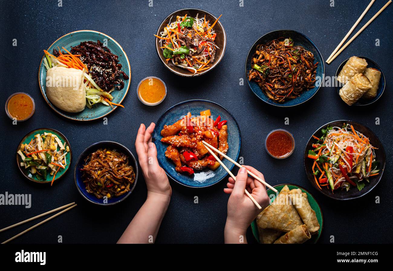 Ensemble de plats chinois sur table, mains de femmes tenant des baguettes : poulet doux et aigre, rouleaux de printemps frits, nouilles, riz, petits pains cuits à la vapeur avec barbecue Banque D'Images