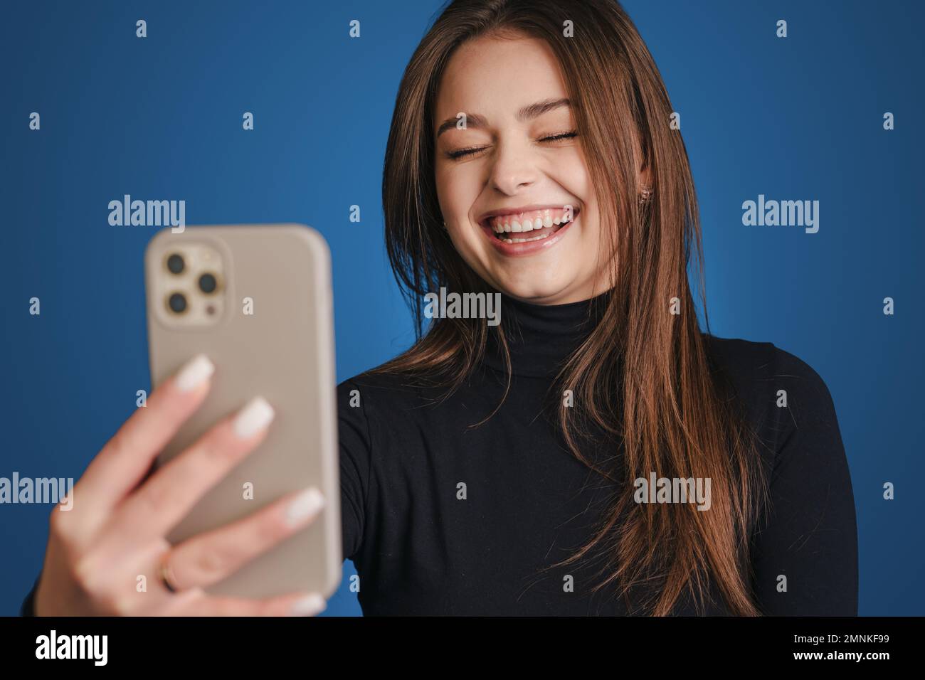 Portrait d'une jeune femme charmante tirant doucement la main avec un smartphone près du visage prenant la photo sur fond bleu. Banque D'Images