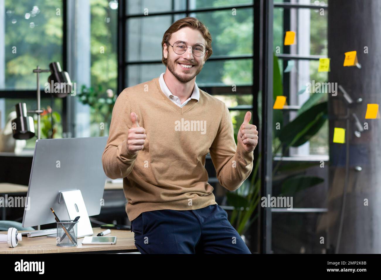 Portrait d'un homme blond heureux à l'intérieur du bureau, d'un homme en vêtements décontractés souriant et regardant l'appareil photo, d'un jeune homme d'affaires réussi montrant les pouces à l'appareil photo, d'un programmeur au travail avec un ordinateur. Banque D'Images