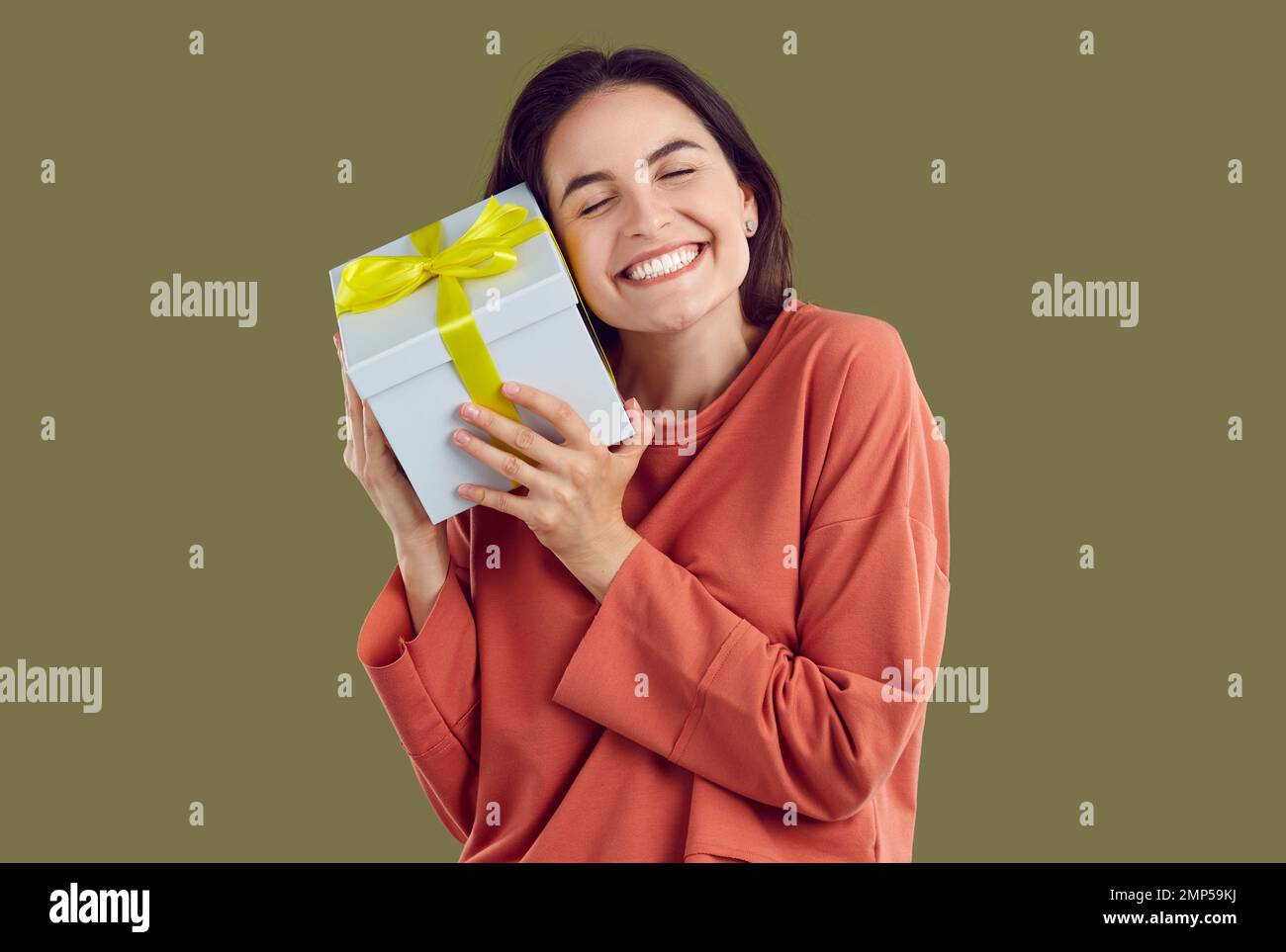 Une femme brune souriante en sweat-shirt rouge embrasse un cadeau avec des yeux fermés sur fond kaki. Banque D'Images