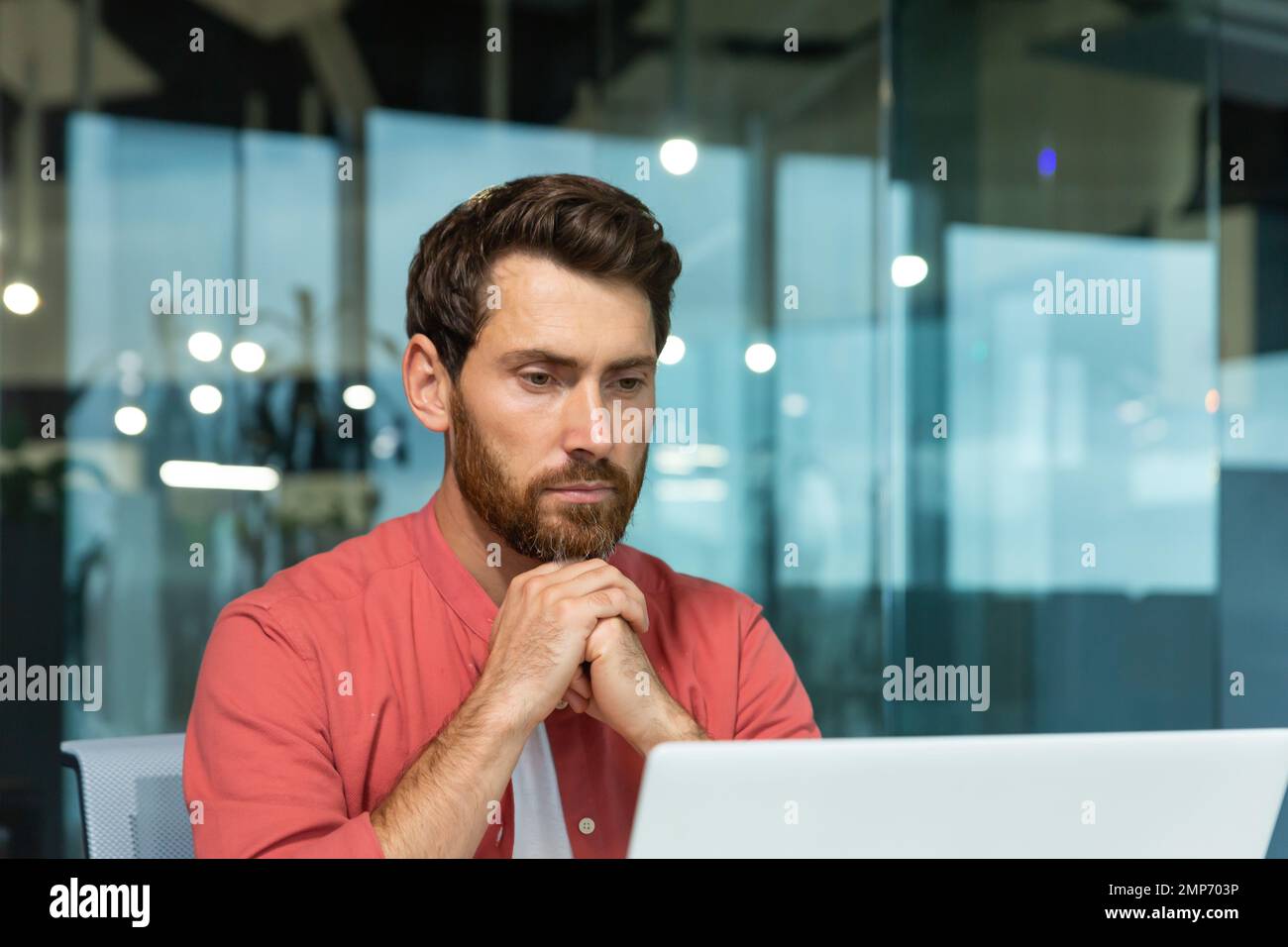 Gros plan. Un jeune homme concentré et réfléchi dans une chemise rouge travaille, des études sur un ordinateur portable au bureau Banque D'Images