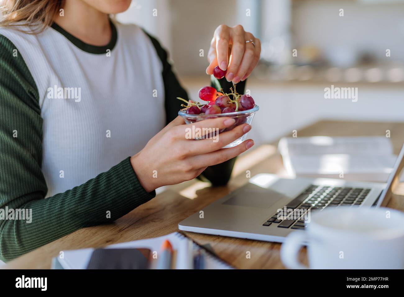 Vue latérale d'une femme tenant un bol avec des raisins au-dessus du bureau avec ordinateur, agenda et smartphone. Concept d'équilibre travail-vie privée. Banque D'Images
