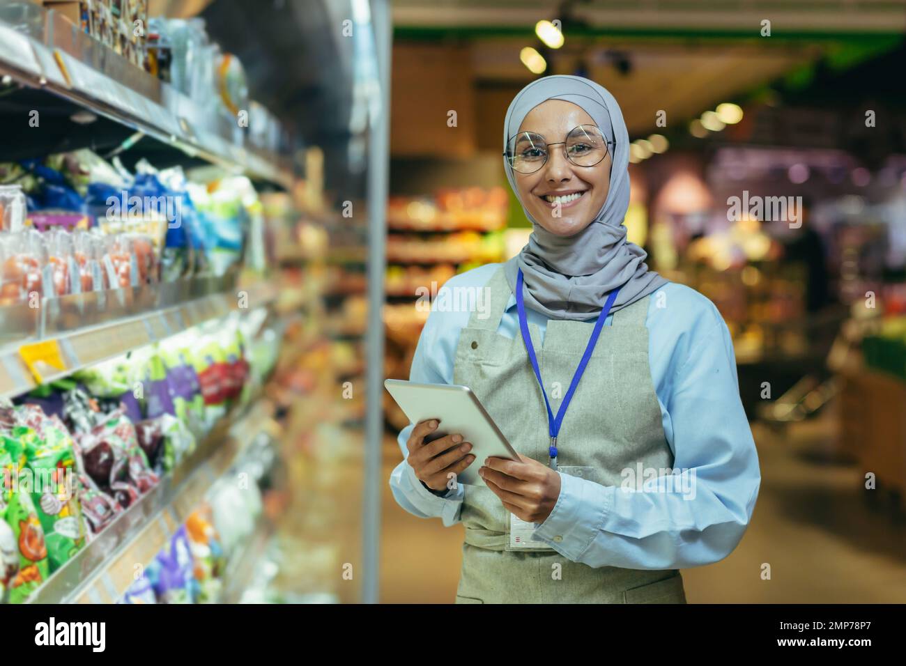 Une jeune femme arabe dans un hijab travaille dans un supermarché, vendeur, consultant, gestionnaire. Debout avec un dossier dans ses mains et un badge dans le rayon épicerie. Il regarde la caméra, sourit. Banque D'Images
