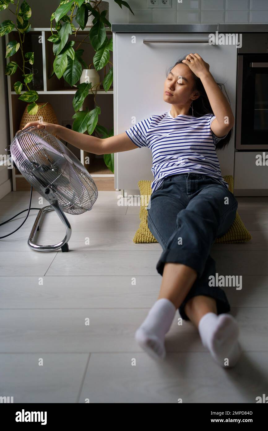 Une jeune femme asiatique décontractée se trouve sur le sol, près du ventilateur de travail et bénéficie de la fraîcheur et de l'air frais Banque D'Images