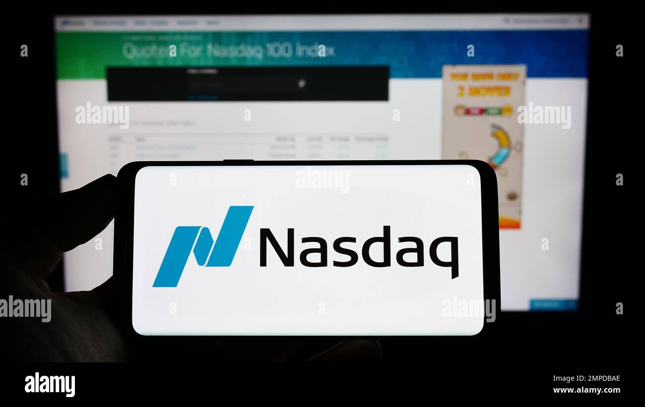 Personne tenant le téléphone portable avec le logo de la bourse américaine Nasdaq sur l'écran en face de la page web d'affaires. Mise au point sur l'affichage du téléphone. Banque D'Images