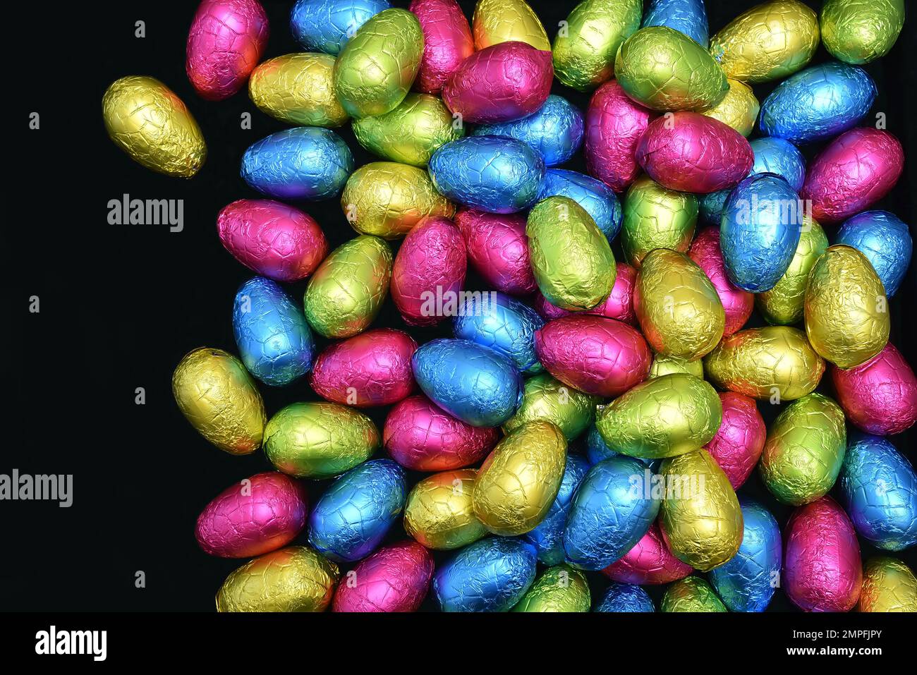Pile de plusieurs couleurs et de différentes tailles d'oeufs de pâques au chocolat enveloppés de papier d'aluminium dans le rose, le bleu, le jaune et le vert lime sur un fond noir. Banque D'Images