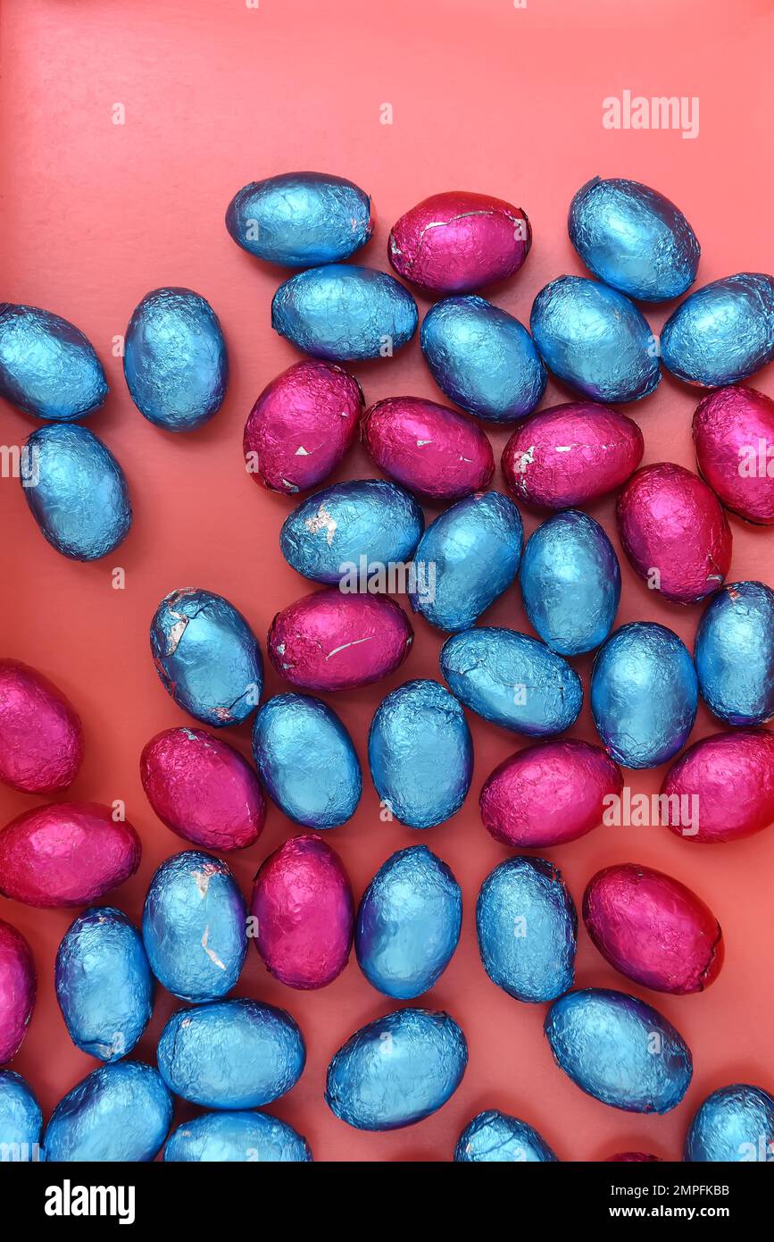 Petits et grands œufs de pâques colorés roses, et bleus enveloppés de papier d'aluminium au chocolat, sur fond orange pêche. Banque D'Images
