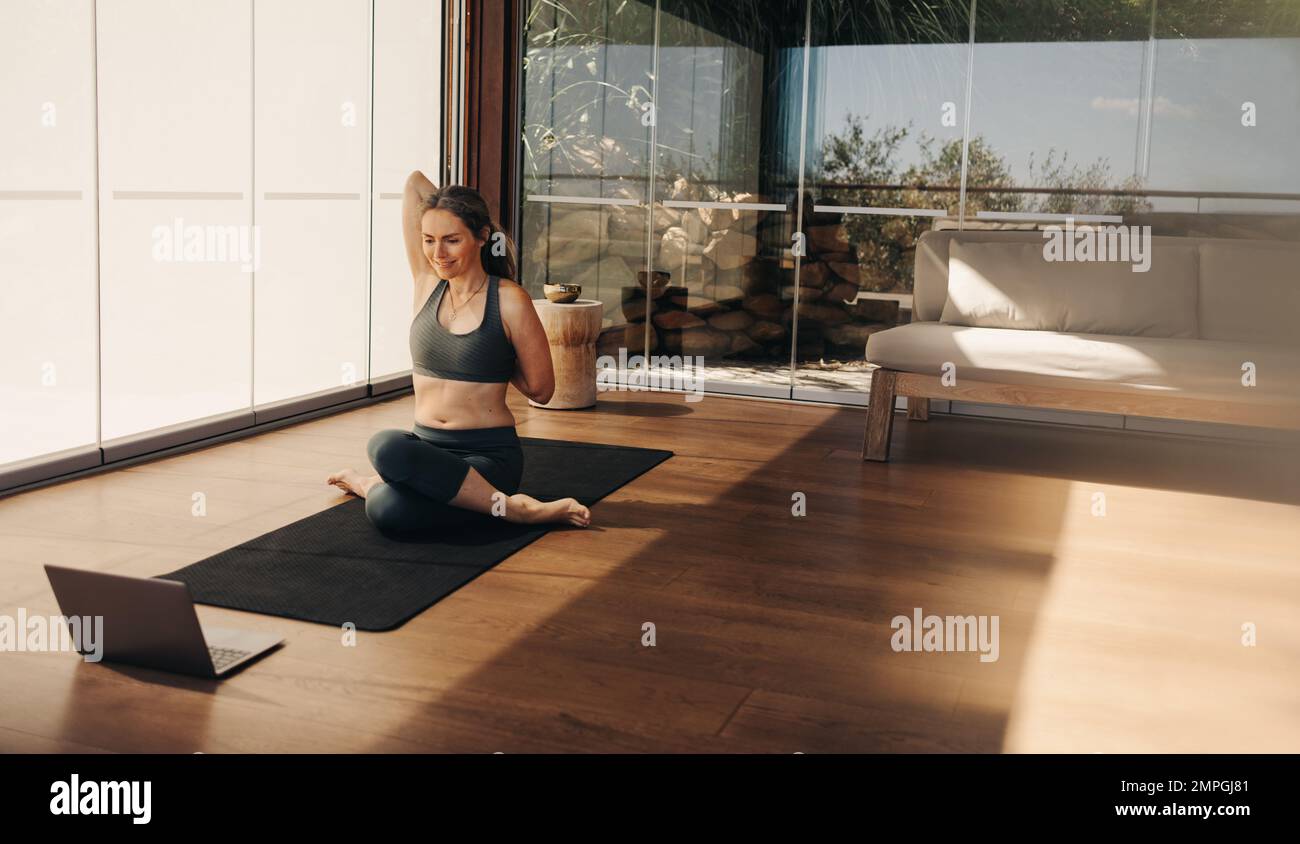 Femme aînée souriante faisant une pose de tête de vache lors d'un cours virtuel de yoga à la maison. Femme mature pratiquant une asana d'étirement sur un tapis de yoga. Femme heureuse Banque D'Images