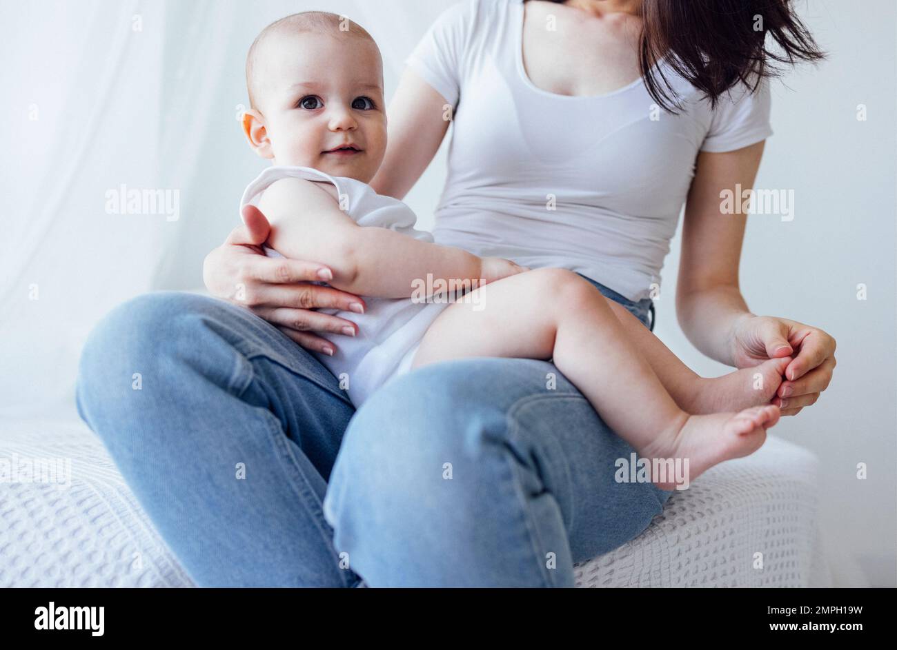 Jeune belle mère embrassant son bébé mignon sur la joue. La brune blanche touche son enfant souriant avec son visage. Joli bébé dans un body blanc avec Banque D'Images