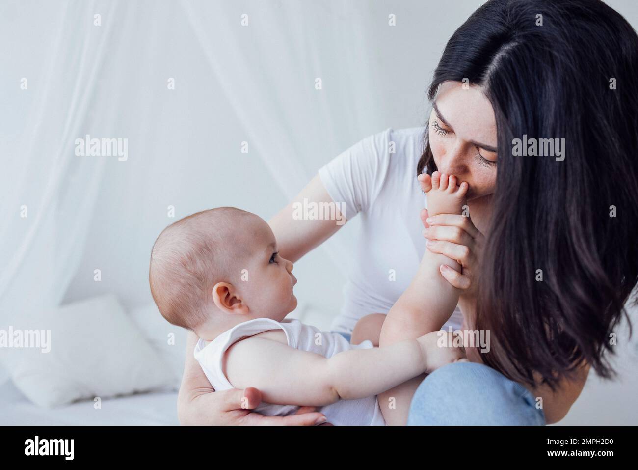 Jeune belle mère embrasse son bébé mignon sur la joue. La brune blanche touche son enfant souriant avec son visage. Joli bébé dans un body blanc avec h Banque D'Images