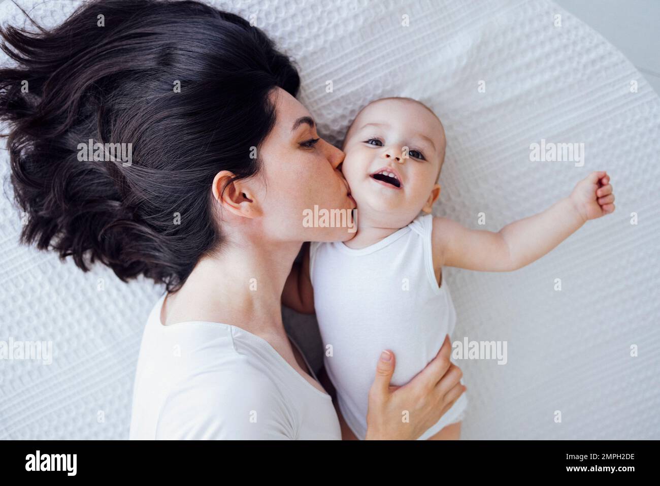 Jeune belle mère embrasse son bébé mignon sur la joue. La brune blanche touche son enfant souriant avec son visage. Joli bébé dans un body blanc avec h Banque D'Images