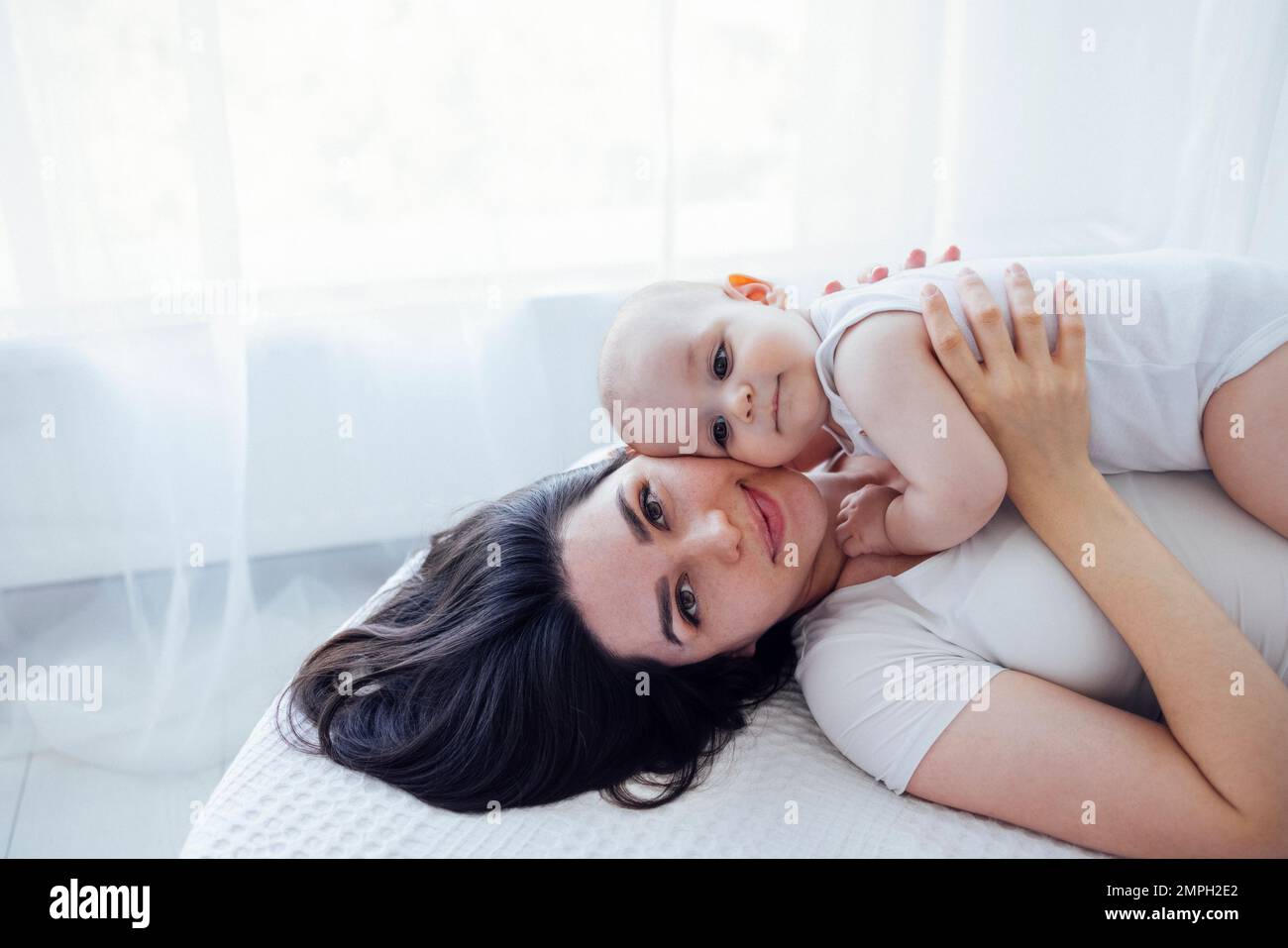 Jeune belle mère embrassant son bébé mignon sur la joue. La brune blanche touche son enfant souriant avec son visage. Joli bébé dans un body blanc avec Banque D'Images