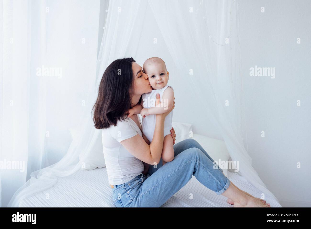 Jeune belle mère embrasse son bébé mignon sur la joue. La brune blanche touche son enfant souriant avec son visage. Joli bébé dans un body blanc avec h Banque D'Images