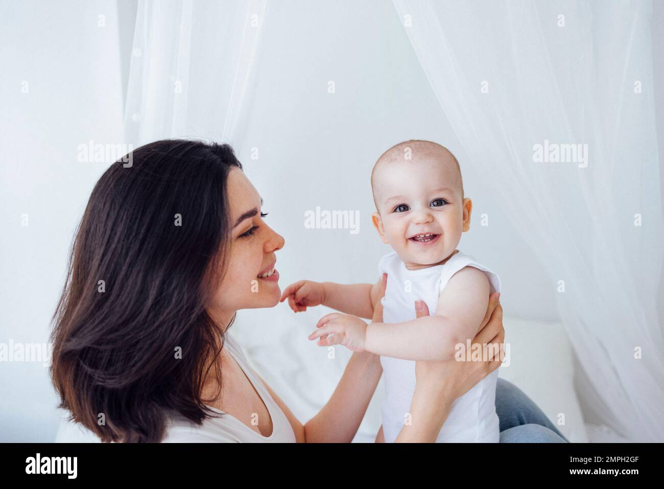 Jeune belle mère embrassant son bébé mignon sur la joue. La brune blanche touche son enfant souriant avec son visage. Joli bébé dans un body blanc avec Banque D'Images