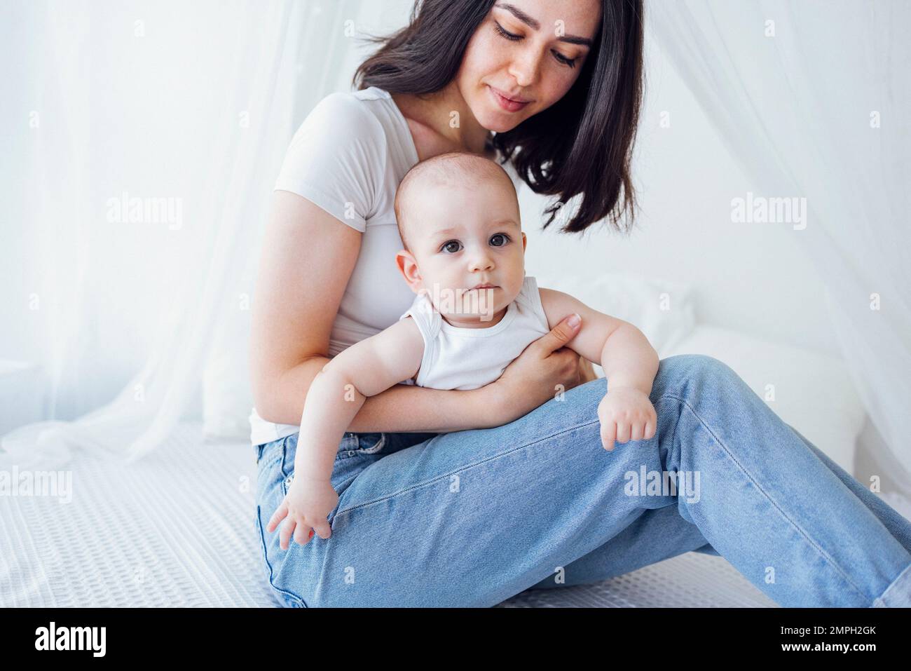 Jeune belle mère embrassant son bébé mignon sur la joue. La brune blanche touche son enfant souriant avec son visage. Joli bébé dans un body blanc avec Banque D'Images