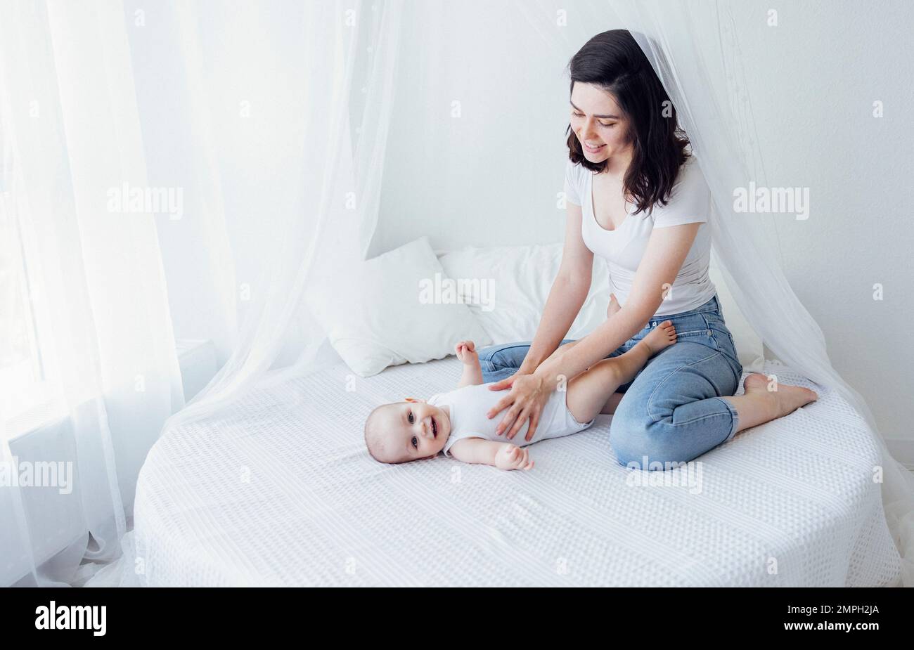 Jeune belle mère embrassant son bébé mignon sur la joue. La brune blanche touche son enfant souriant avec son visage. Joli bébé dans un body blanc avec Banque D'Images