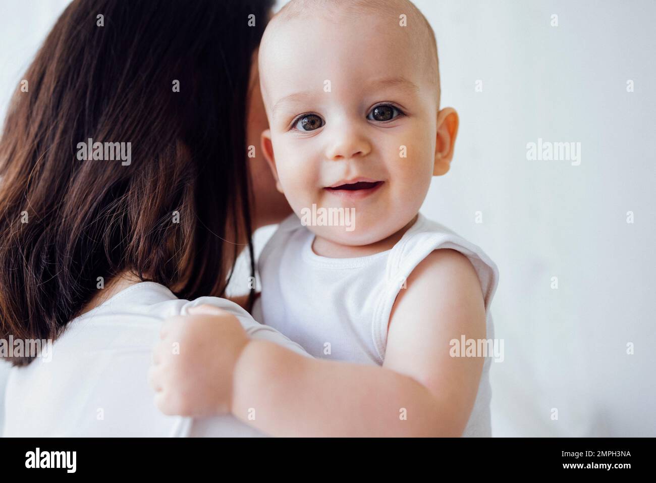 Jeune belle mère embrassant son bébé mignon sur la joue. La brune blanche touche son enfant souriant avec son visage. Joli bébé dans un body blanc avec Banque D'Images