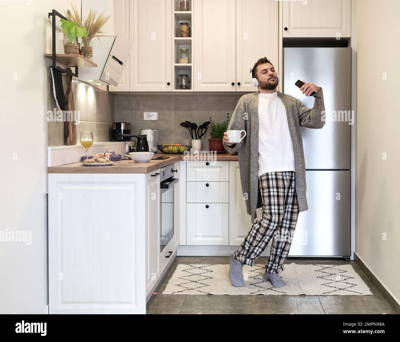 Homme barbu adulte avec un casque tenant une tasse à café et un téléphone portable dans ses mains dansant et appréciant la musique avec les yeux fermés dans la cuisine domestique dans Banque D'Images