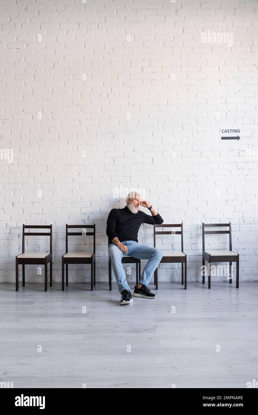 homme aux cheveux gris lassés assis sur une chaise près du mur blanc et attendant la coulée Banque D'Images