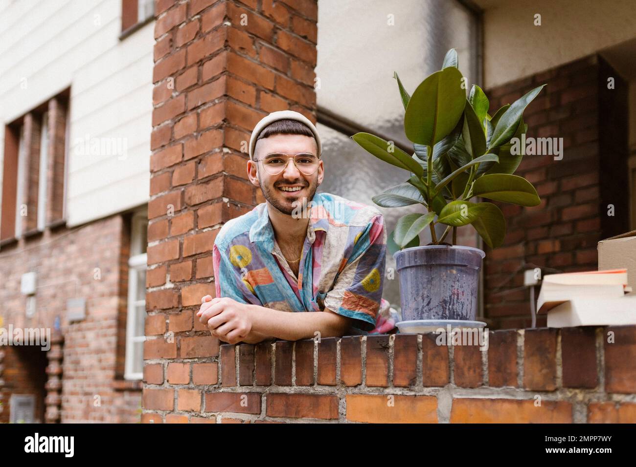 Portrait d'un homme souriant penché par une plante en pot sur un mur de briques Banque D'Images