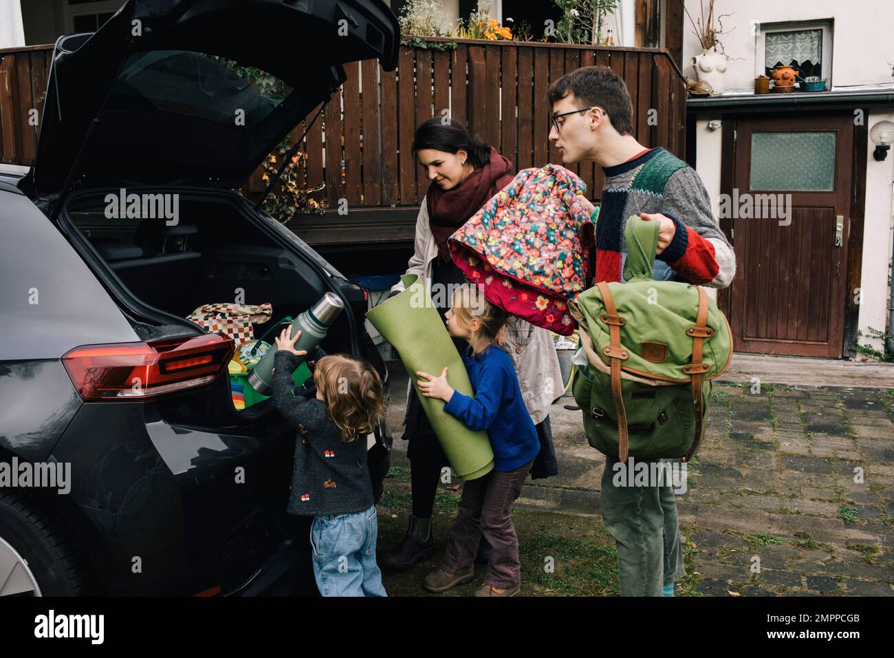 Une famille excitée se charge des produits de pique-nique dans le coffre de voiture électrique à l'extérieur de la maison Banque D'Images