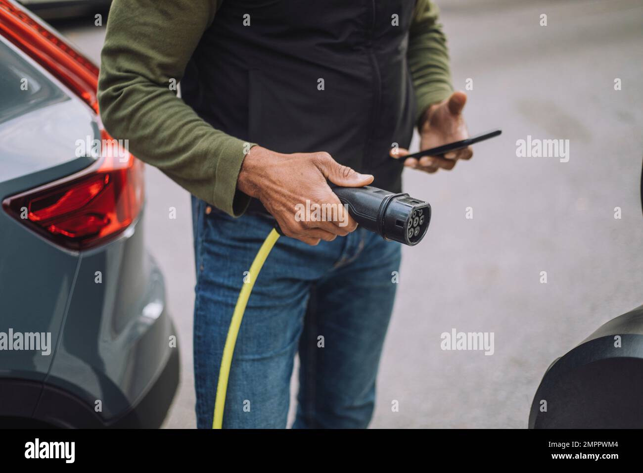 Section médiane de l'homme mature tenant le smartphone et la fiche du chargeur électrique à la station Banque D'Images