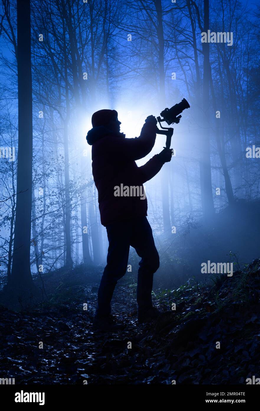 Silhouette d'un vidéaste dans une forêt brumeuse, rétro-éclairé de façon spectaculaire avec une couleur bleu froid et des rayons de lumière derrière lui Banque D'Images