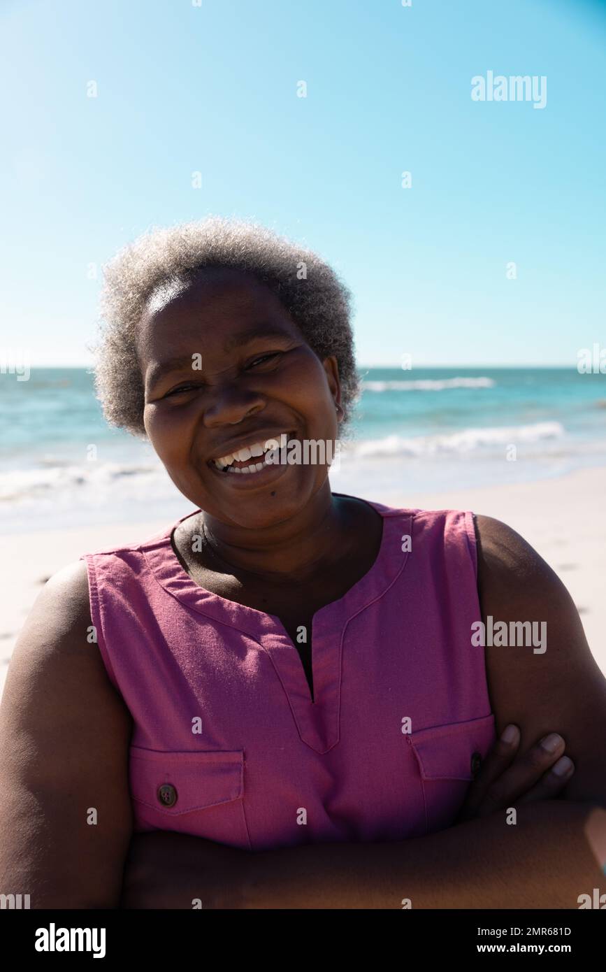 Joyeuse femme afro-américaine âgée avec les armes croisées en profitant de vacances à la plage sous ciel clair Banque D'Images