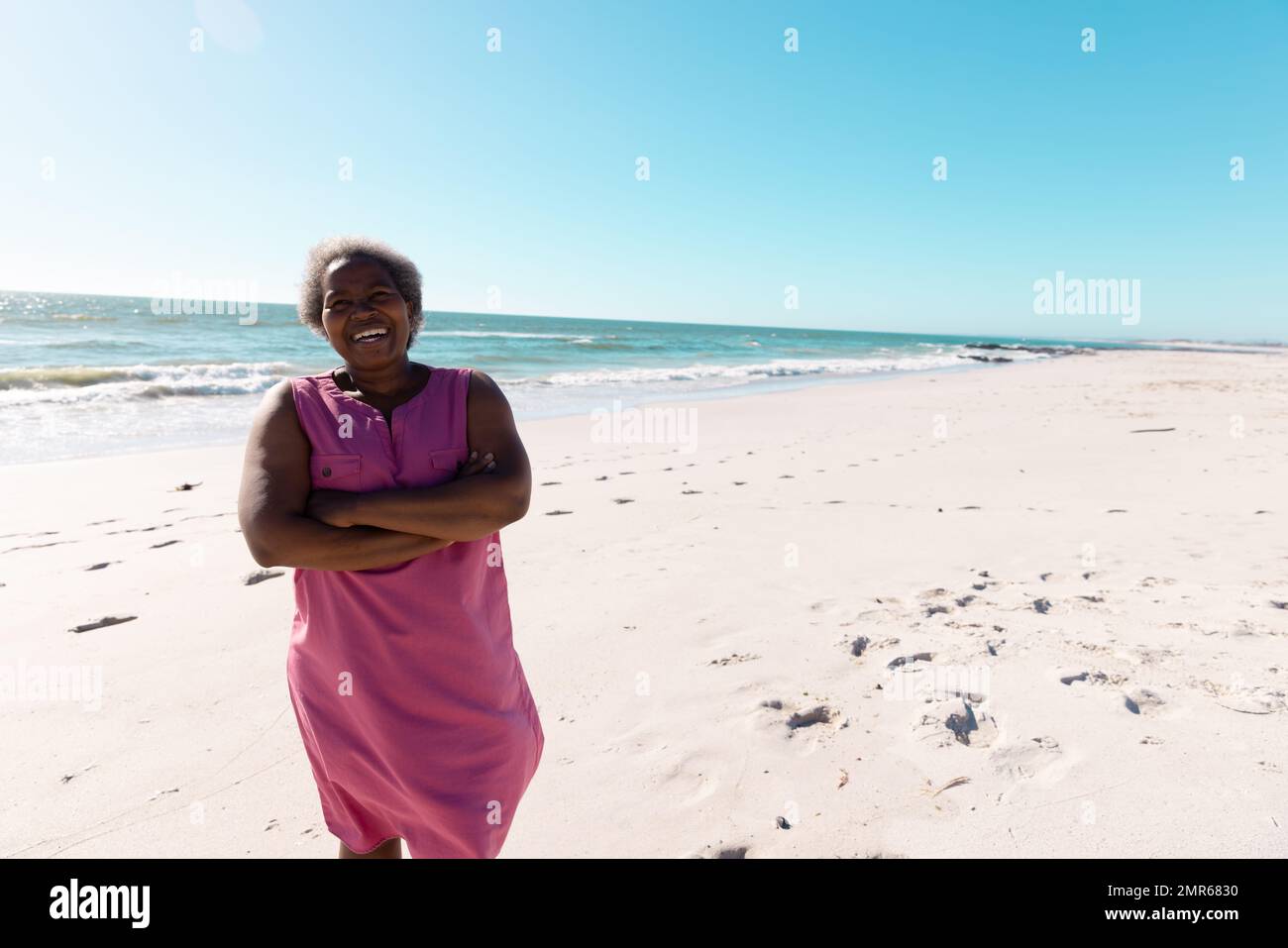Femme afro-américaine âgée avec des armes croisées debout sur une plage de sable sous un ciel clair le jour ensoleillé Banque D'Images