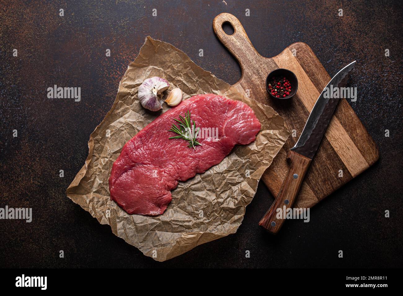 Filet de bœuf cru maigre steak sur du papier à pâtisserie avec romarin, ail et épices sur fond de béton rustique brun foncé avec couteau de dessus plat, d Banque D'Images