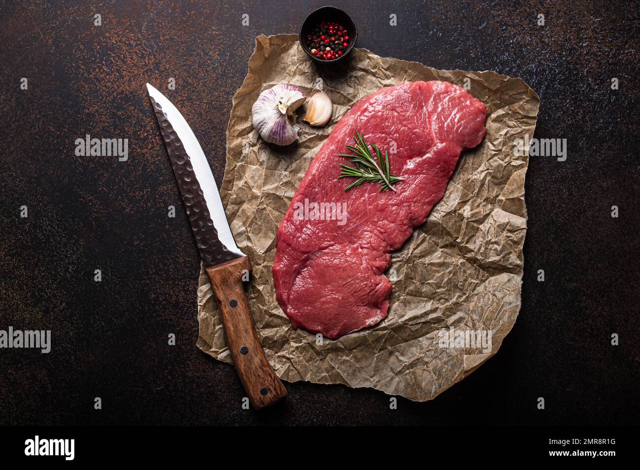 Filet de bœuf cru maigre steak sur du papier à pâtisserie avec romarin, ail et épices sur fond de béton rustique brun foncé avec couteau de dessus plat, d Banque D'Images