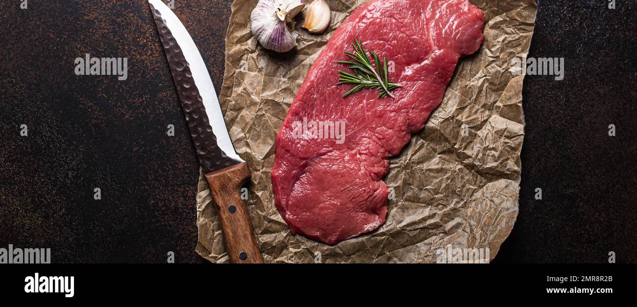 Filet de bœuf cru maigre steak sur du papier à pâtisserie avec romarin, ail et épices sur fond de béton rustique brun foncé avec couteau de dessus plat, d Banque D'Images