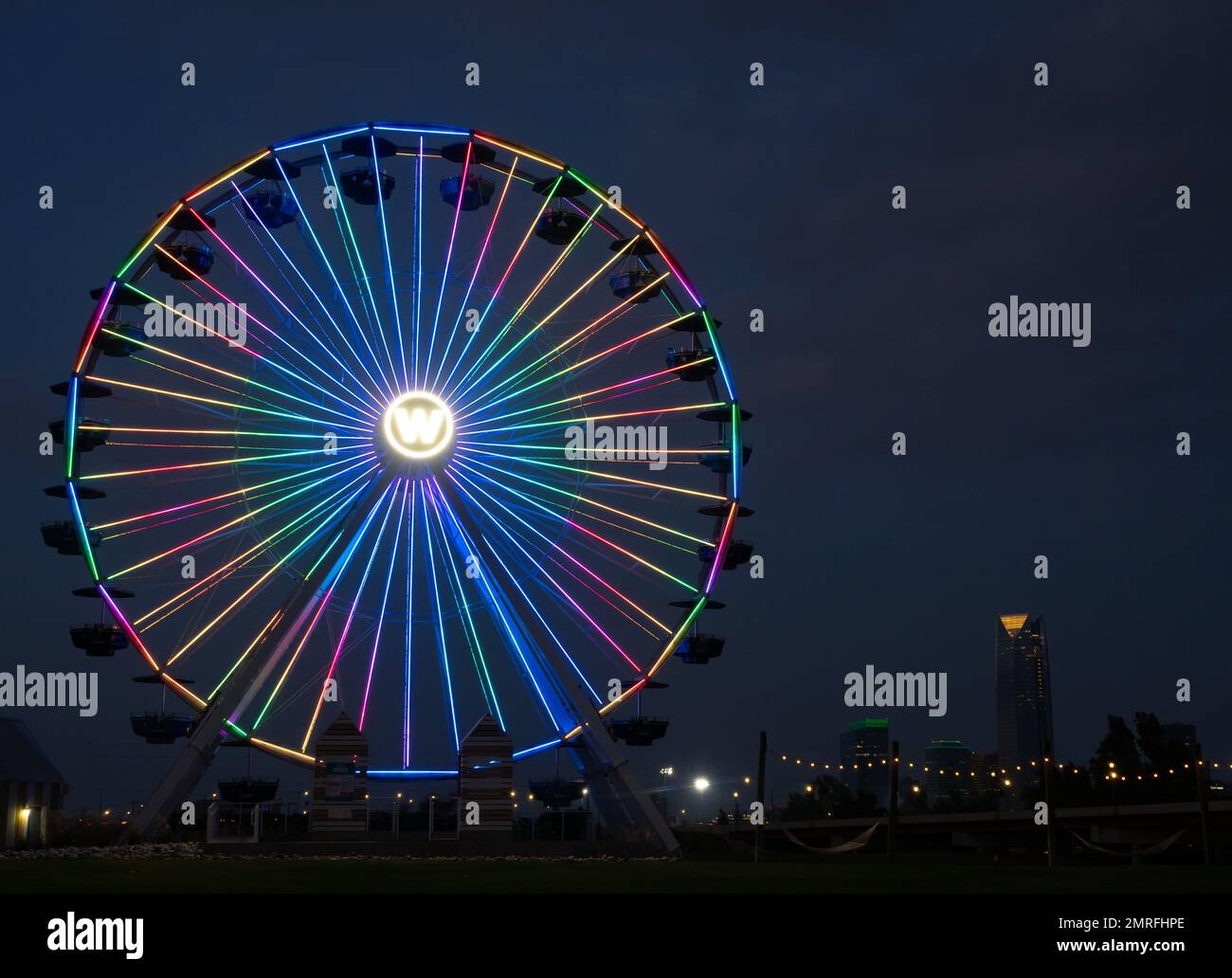 Roue fixe Wheeler Park Ferris éclairée par des néons de couleur arc-en-ciel et photographiée la nuit. Le centre-ville d'Oklahoma City est en arrière-plan. Banque D'Images