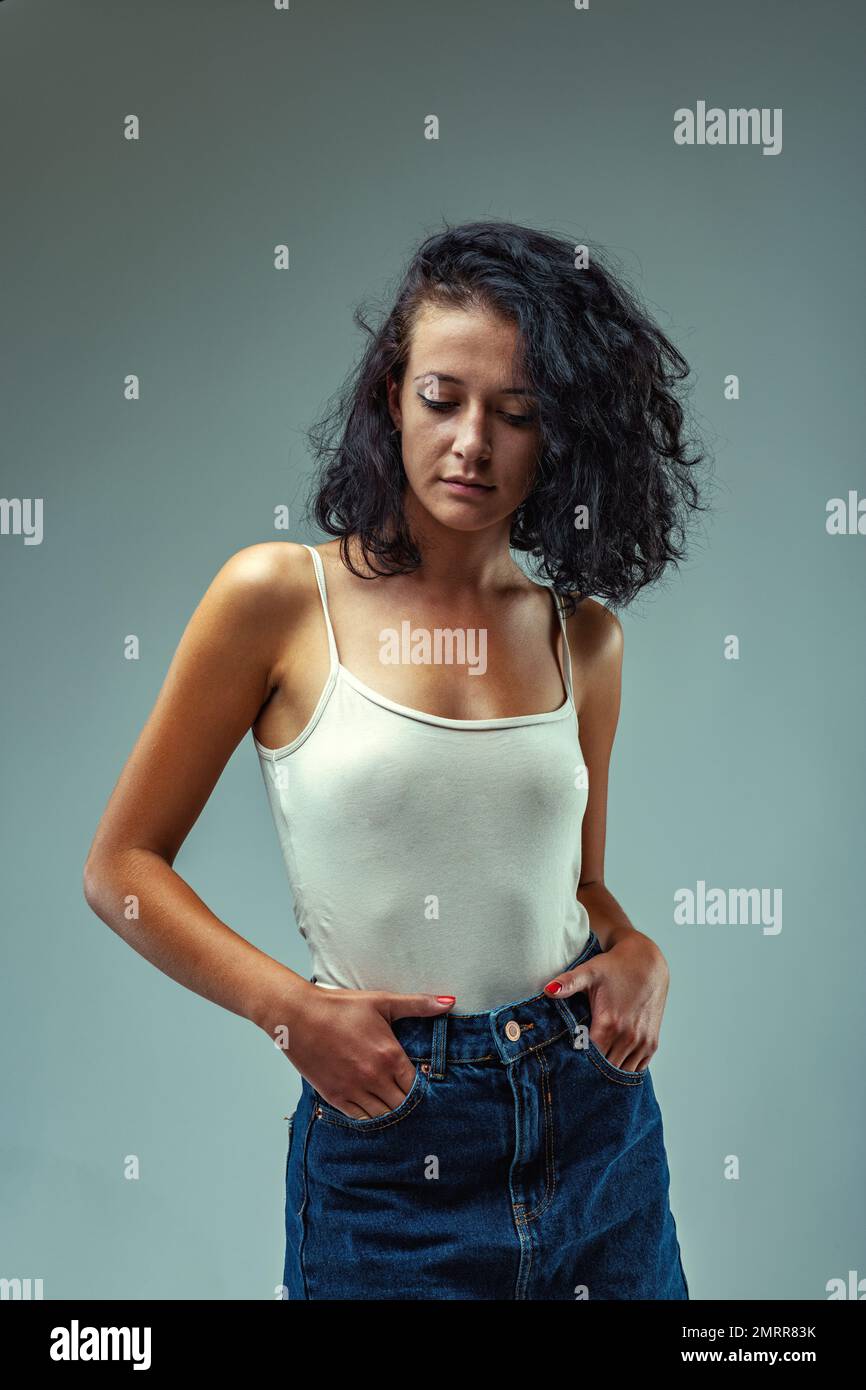 Fille triste ou pensive debout dans un débardeur blanc et jupe jean tient les mains dans les poches tout en gardant les yeux vers le bas. Bien qu'elle soit très belle et attra Banque D'Images