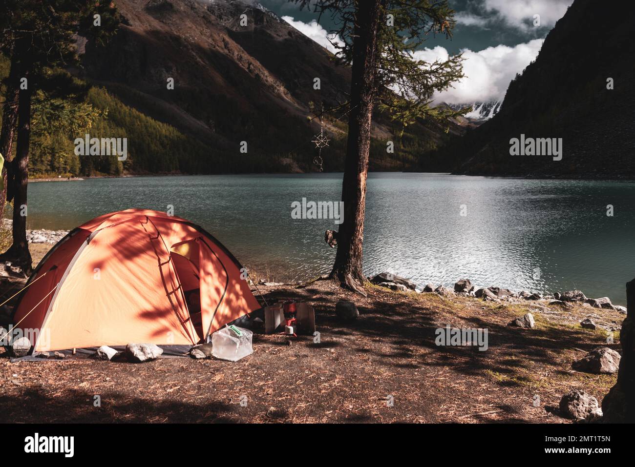 Une tente orange se dresse dans les arbres sur la rive du lac turquoise de haute altitude Shavlinskoye en Altai dans l'après-midi après une pluie. Banque D'Images