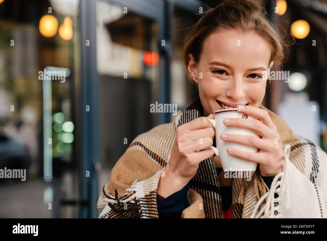 Joyeuse jeune femme blonde dans le tissu écossais chaud boire du thé chaud tout en se promenant dehors dans la rue de la ville Banque D'Images