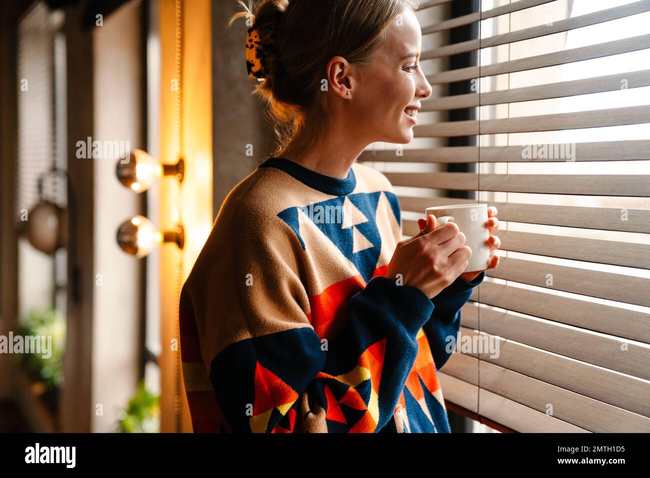 Jeune belle femme souriante portant un chandail coloré regardant par la fenêtre tout en buvant un café dans le café Banque D'Images