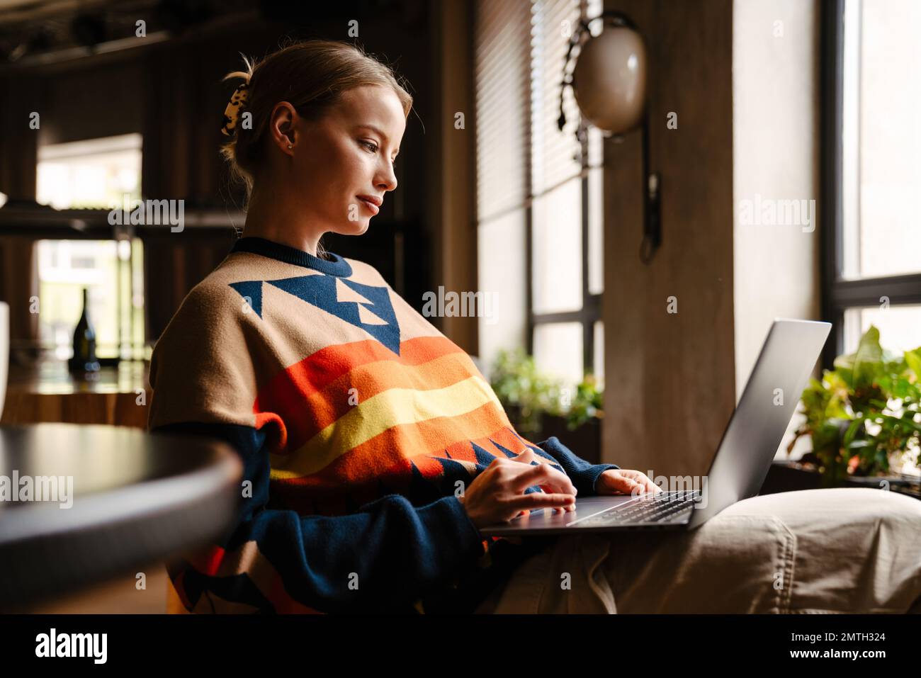 Jeune belle femme portant un chandail coloré travaillant sur un ordinateur portable dans un café Banque D'Images