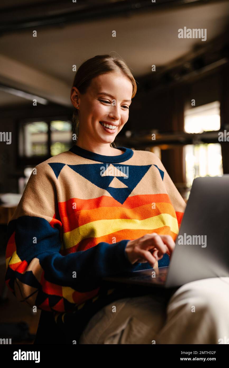 Jeune belle femme souriante portant un chandail coloré travaillant sur un ordinateur portable dans un café Banque D'Images