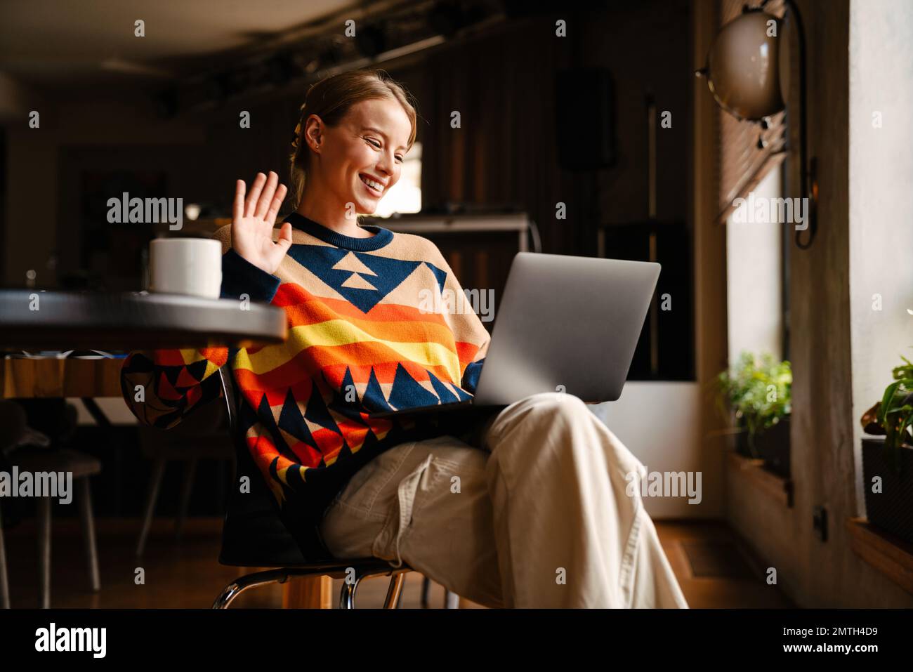 Jeune belle femme souriante portant un chandail coloré ayant des appels vidéo par ordinateur portable et agitant tout en étant assis dans un café Banque D'Images