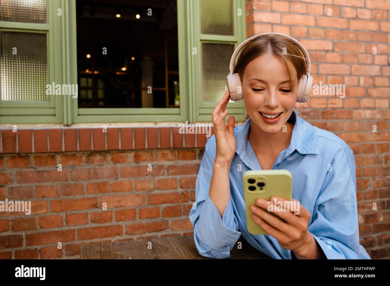 Portrait d'une jeune femme heureuse souriante qui touche son casque sans fil, tient et utilise son téléphone, tout en étant assise près de la fenêtre à l'extérieur Banque D'Images