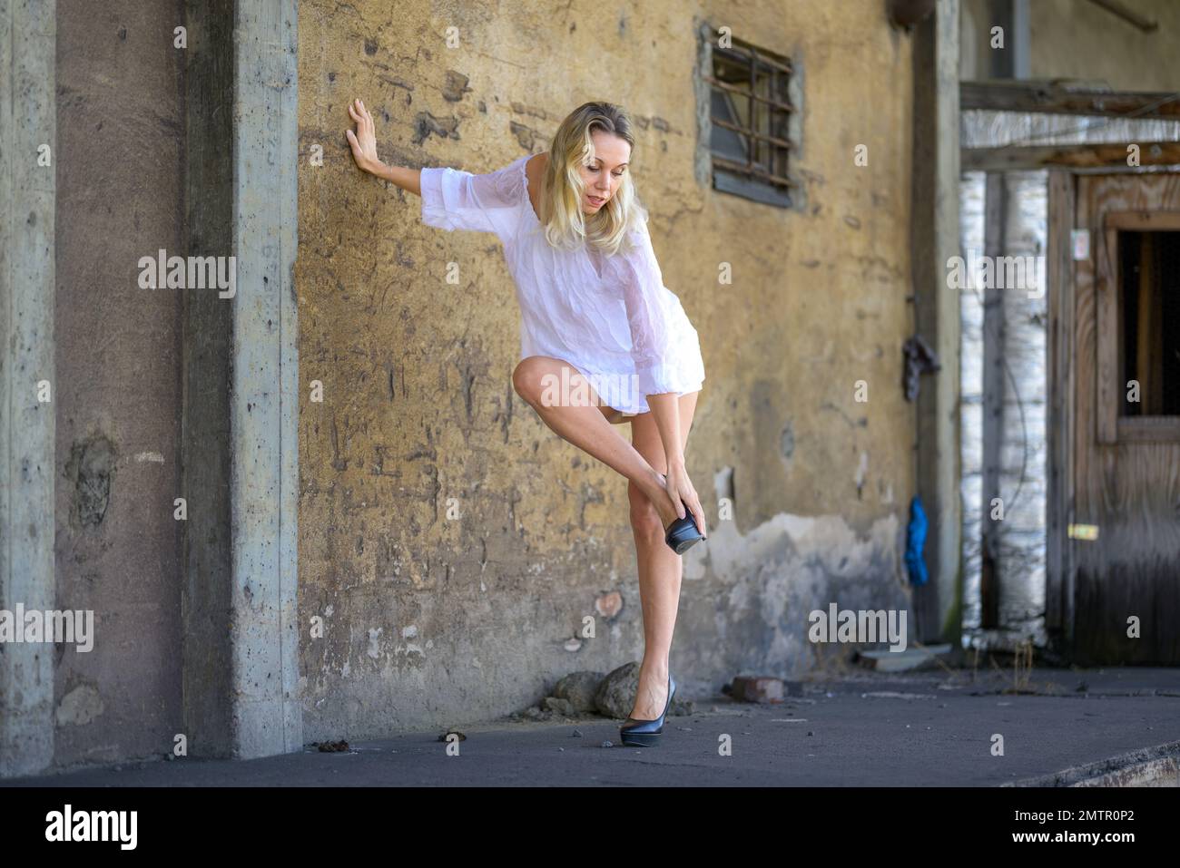 Blonde d'âge moyen belle femme portant une robe courte blanche penchée contre un mur mettant ses chaussures Banque D'Images