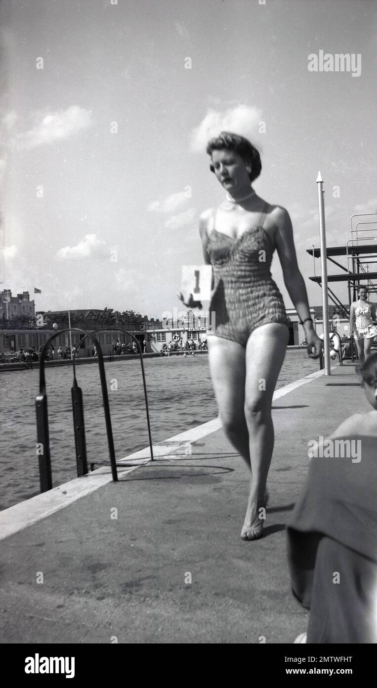 1950s, historique, une grande femme élégante candidat dans son maillot de bain, portant le no 1 sur une carte, marchant au bord d'une piscine, prenant part à un concours de beauté de bord de mer, Skegness, Angleterre, Royaume-Uni. Vu au loin, le Pier Pavilion. Banque D'Images