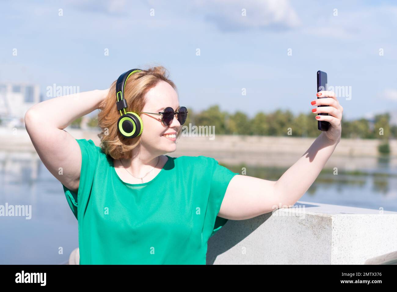 Une femme heureuse dans des lunettes de soleil et des écouteurs prenant une photo de selfie au téléphone, discutant avec des amis dans la rue. Formation à distance, formation en ligne et Banque D'Images