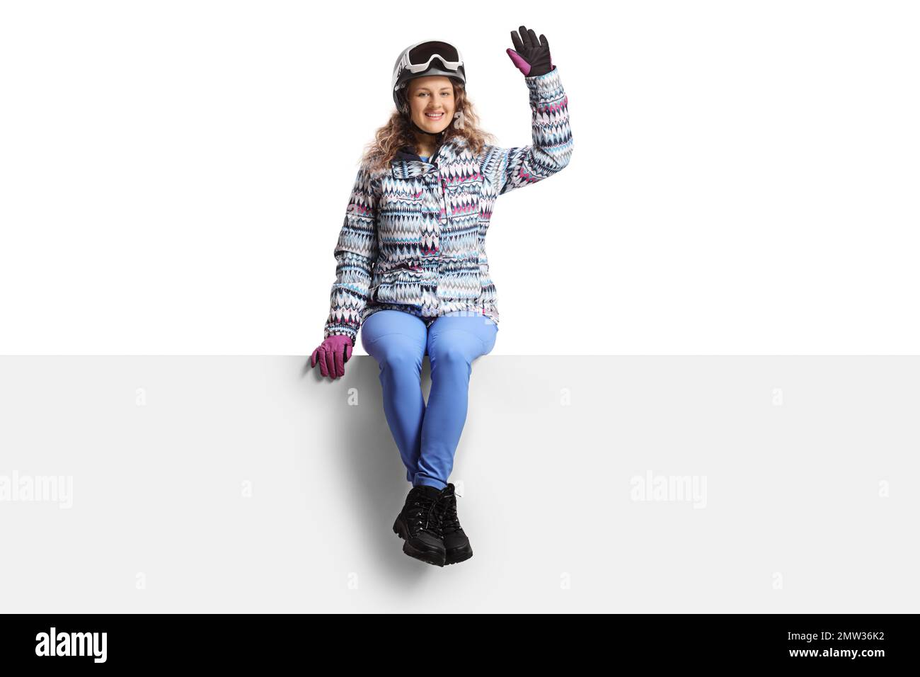 Jeune femme portant un équipement de ski assise sur un panneau vierge et agitant isolée sur fond blanc Banque D'Images