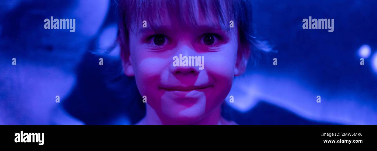 portrait visage mignon heureux candié caucasien petit garçon de six ans avec de longs cheveux blonds dans le studio éclairé par lumière bleue violet néon. banne Banque D'Images